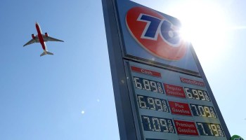 A plane flies behind a sign advertising gas prices over $6 a gallon.