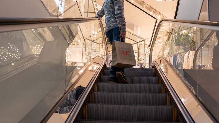 A person on an escalator holds an H&M bag. They are wearing blue jeans and a gray pullover.