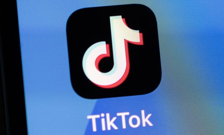 The TikTok app icon — a white musical note with a flag on a black background — on a smartphone.
