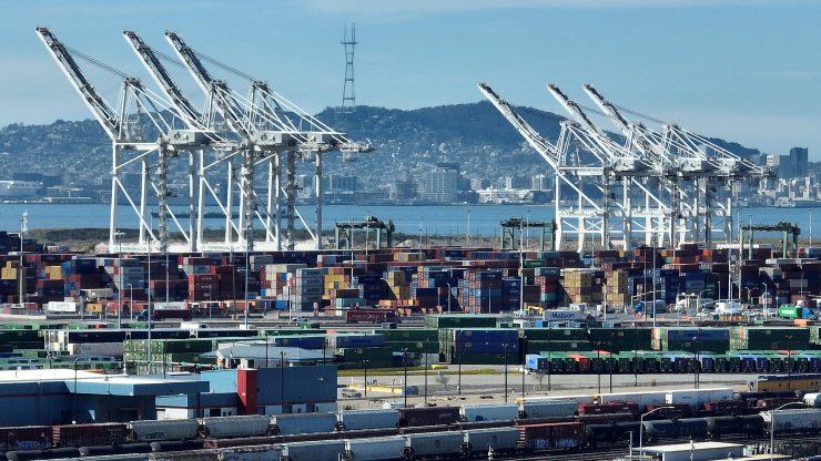 Rail cars with shipping containers pass in front of dozens of stacked shipping containers at the Port of Oakland in California. Behind the stacks are massive white cranes, and behind that is the port, water and a hillside dotted with buildings.