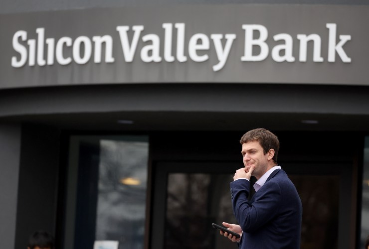 A person in a dark suit, one hand on his chin and the other holding a phone, stands outside of the shuttered Silicon Valley Bank headquarters.