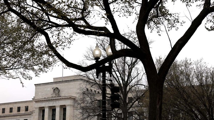 The facade of the Federal Reserve building is seen behind blooming tree branches and a cloudy sky.