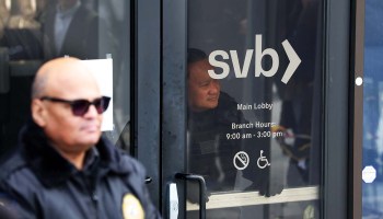 A security guard in a black jacket and dark sunglasses looks out in front of the doors of a Silicon Valley Bank location. Behind SVB's logo, another man looks out.