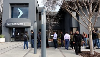 A line of people in front on a concrete sidewalk in front of a modern, gray building with the Silicon Valley Bank logo, a right-pointing arrow or caret.
