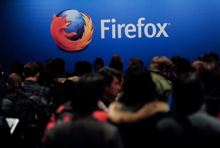 A group of people at a conference sit before a large photo of the Firefox web browser logo — a red fox wrapped around planet Earth, its tail turning into orange flames.