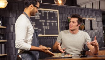A customer in a grey shirt with spikey black hair and long sideburns looks disgusted and disgruntled while pointing toward a plate of food while a bearded waiter wearing a white shirt and grey apron stands nearby, pointing toward the food.