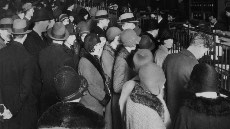 In a black-and-white photo from 1929, a crowd of men in suits and Fedoras and women dressed in cloche hats and fur-trimmed jackets wait outside a gate to withdraw their money.