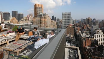 People sit and work on a rooftop terrace. The New York City skyline is seen in the background.