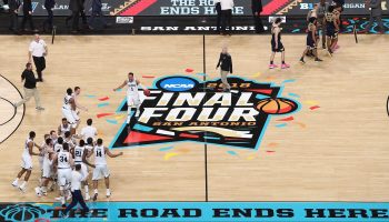 On a light wood basketball court, players run up and down chasing a basketball. There is a massive Final Four logo painted in the center of the court.