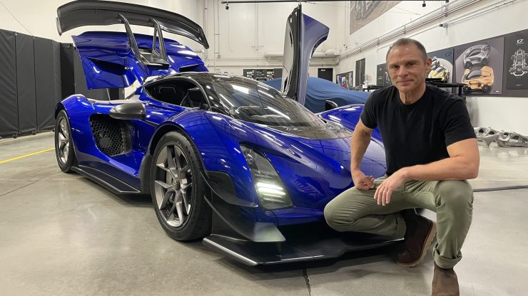 Kevin Czinger, a man with cropped hair wearing a black T-shirt and green pants, kneels next to a sleek, futuristic blue car — the Czinger 21C.