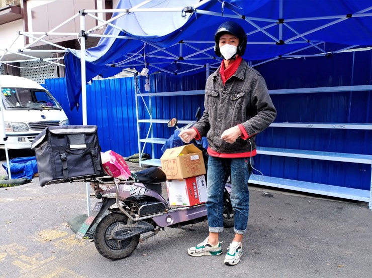 Il corriere Wang Xiu indossa una maschera e indossa blue jeans, una maglietta rossa e una giacca di jeans nera.  È in piedi accanto alla sua bici elettrica. 