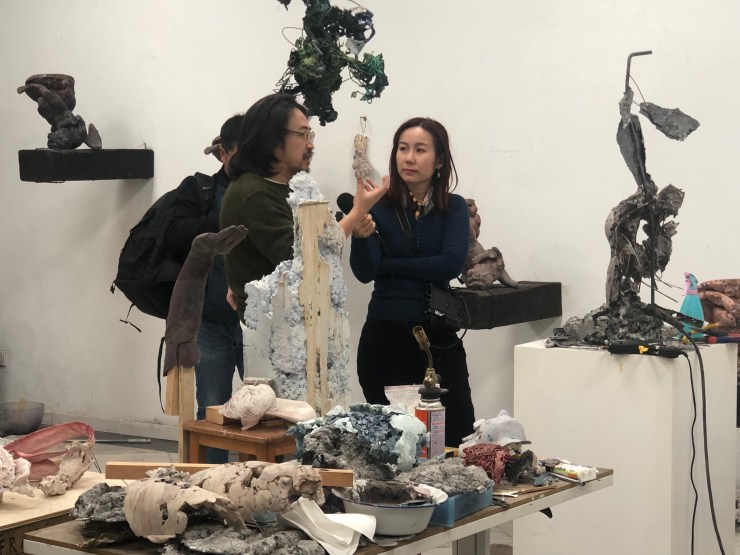 Marketplace's Jennifer Pak wears all black. She is holding a microphone pointed toward Jin Shan, who holds his hand up and speaks into the microphone. They are in Shan's Shanghai studio and are surrounded by modern scultpures. 