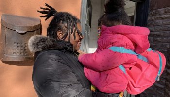 Mansie Meikle wears a black parka with her hair up. She is holding her 4-year-old daughter, who is also wearing her hair up and has on a cute pink coat and little backback.