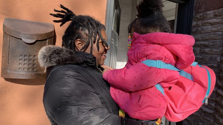 Mansie Meikle wears a black parka with her hair up. She is holding her 4-year-old daughter, who is also wearing her hair up and has on a cute pink coat and little backback.
