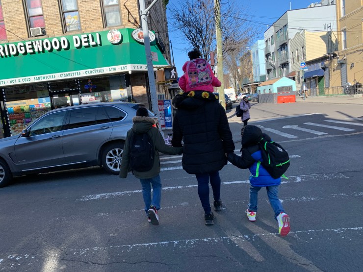 A mom walks hand-in-hand with two of her kids while one is sitting on her shoulders. They are all bundled up, wearing winter coats and walk across a street in New York City.