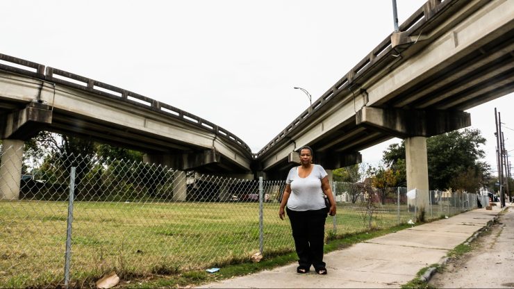 Amy Stelly, a woman with short hair, a gray t-shirt and dark blue jeans, stands in front of a fenced off grassy patch where two portions of an elevated highway come together.