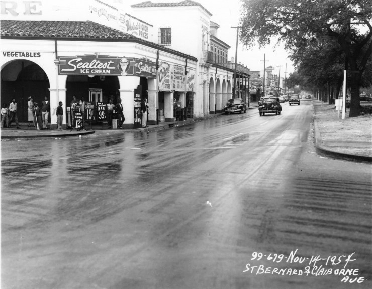 A black and white photo shows The Circle Food Market, a white building with clay tile roof. It is a market that advertises vegetables and ice cream. People stand near its entrance. On the other side of the road, Clairborne Avenue, mature trees stand tall as cars drive by.
