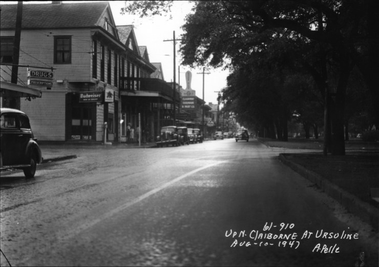 A black and white photo shows Clairborne Avenue in the 1940s. On the left side of the road, two-story businesses and homes are flanked by parked automobiles. On the right side are large, mature trees that shade the street.
