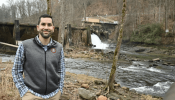Joey Owle wears khaki pants, a blue gingham shirt and gray vest. Behind him is a dam and flowing water in a forest.