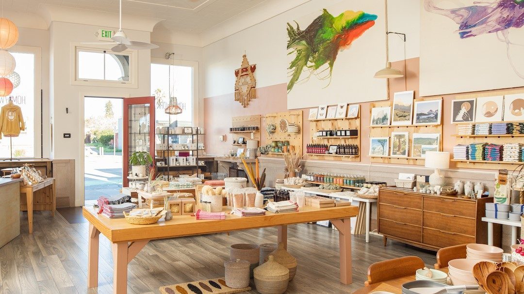 Inside the home/work gift shop, the white walls are covered in artwork and a wooden table displays linens, plates and other tableware.  Baskets are placed under the table.