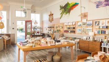 Inside Home/Work gift shop, the white walls are covered in artwork and a wooden table displays linens, dishes and other tableware. Baskets sit under the table.