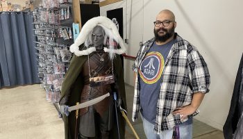 Mark Warren, a tall bald man with a black beard is wearing black glasses, a t-shirt, white and blue flannel and light blue jeans. He is standing next to a Dungeons & Dragons character that is slightly shorter than him. The character is grey, has a sword and is wearing brown armor with a green cape and white hood.