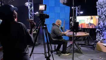A woman, Krenée Tolson, stands at a camera on a tripod. The camera is directed at a man wearing black, Geremy Dixon. He is seated at a table, speaking into a microphone. Another person is seen on a television screen. Tolson and Dixon are in a studio space, with studio lighting and blue acoustic squares.