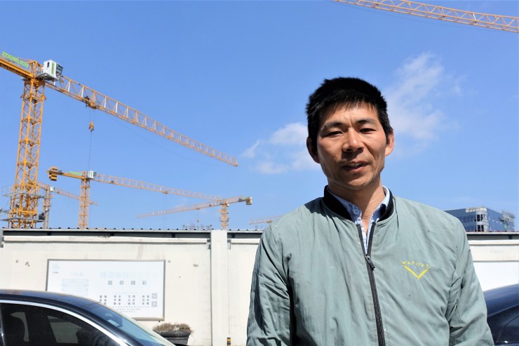 Zhang Xianfeng Wears A Blue And Green Jacket And Stands In Front Of The Construction Site. 