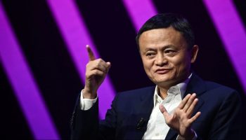 Jack Ma wears a black suit with a white button-up. He is mid-sentence and has both of his hands facing out, one pointing upward. He sits in front of a black and purple striped background.