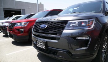 A series of Ford Explorer SUVs — white, red, dark blue — are lined up in a row at a Ford dealership.