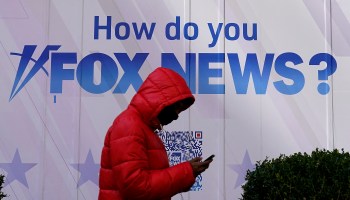 A person wearing a red jacket looks at their phone. Behind them, the Fox News headquarters