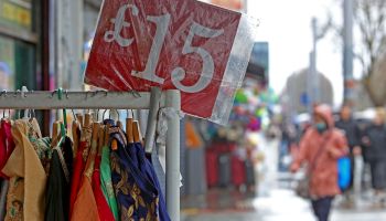 A sign displays the price in pound sterling for clothes at a market. The sign reads 15 pounds and it sits on top of a rack of clothes.