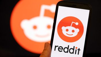 A hand holds a phone with the Reddit logo. An enlarged Reddit logo is seen in the background.