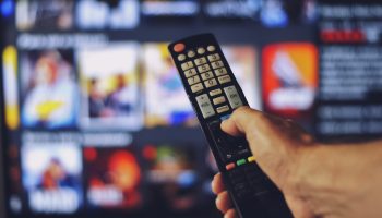 A male hand holding TV remote control. The focus of the photo is on the remote control. The background of several images on TV screen is blurred.