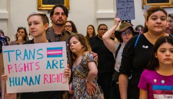 Demonstrators gather at the Texas State Capitol. A family is seen close together. A young person holds a sign that reads "Trans rights are human rights," written in blue and pink.