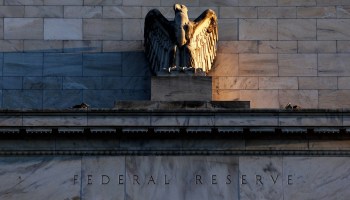 The exterior of the Federal Reserve building is clad in white marble. The statue of a bald eagle sits atop stone that reads "Federal Reserve."