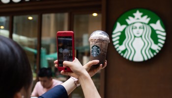 A woman holds up a Starbucks Frappuccino in front of a Starbucks sign and snaps a photo of it.