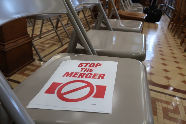 In a row of beige folding chairs, a small placard says "Stop the merger" in red.