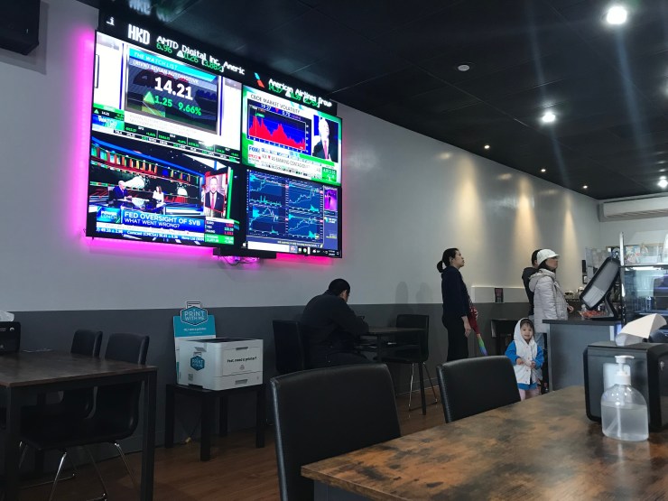 A stock ticker hangs above four TV screens showing financial news and a Twitch live stream at The Bagel Exchange in Alhambra, California.