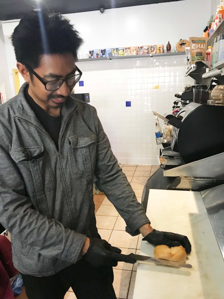 Ryan Natividad is wearing a gray jacket and gloves.  He cuts a bagel in the kitchen of his shop, The Bagel Exchange. 