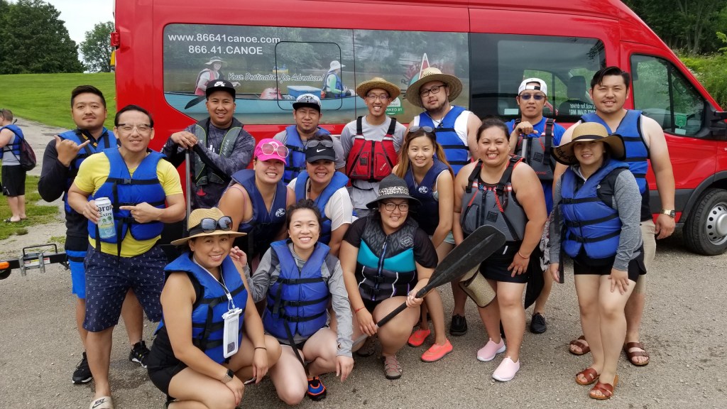 People in life vests stand and smile in front of a bus advertising Teuber's canoeing business.