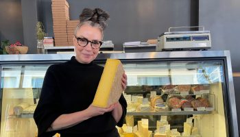Lydia Clarke in front of her "happy place," the cheese case. Clarke has her gray hair up in a bun. She wears black-framed glasses and all black clothing and holds a half wheel of cheese.
