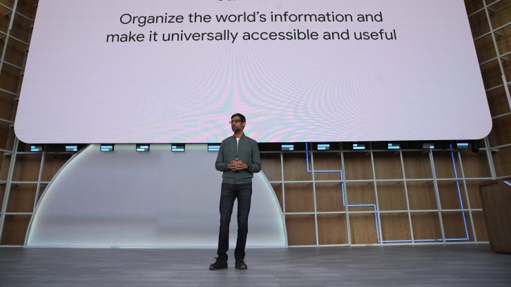 Google CEO Sundar Pichai wears glasses, a gray jacket and blue jeans. He is standing on a stage, speaking. Behind him, a screen reads "Our mission: Organize the world's information and make it universally accessible and useful."