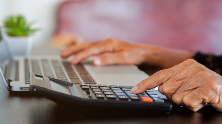 A close-up shows the hands of an older woman. One hand types on a laptop keyboard while they other presses a button on a calculator.