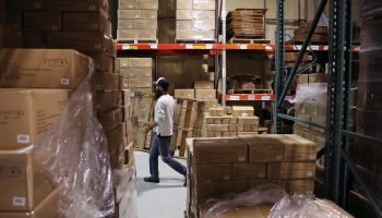 In a warehouse, shelves are lined with pallets full of cardboard boxes. A worker in a white shirt, gray pants and blue baseball cap walks down a row and carries a wooden pallet.