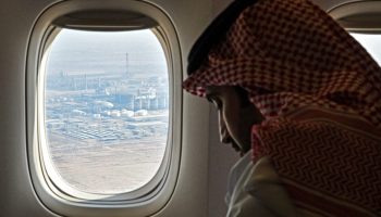 A man with a keffiyeh sits in a plane. Out of the plane window, an oil industrial facility is seen.