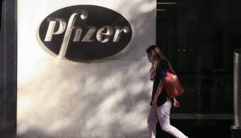 A woman in a face mask walks past a white building with a black Pfizer logo on it.