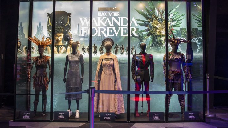 Five mannequins with blank faces wear costumes from "Black Panther: Wakanda Forever." In the center is Ramonda's peach brocade caped gown and regal headpiece.