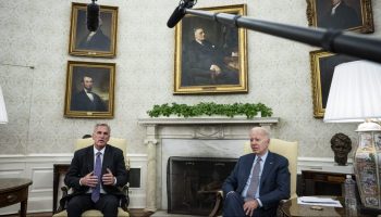 President Joe Biden meets with House Speaker Kevin McCarthy in the Oval Office. They sit in front of a fireplace with paintings of statesmen above them and two large microphones are seen in view.