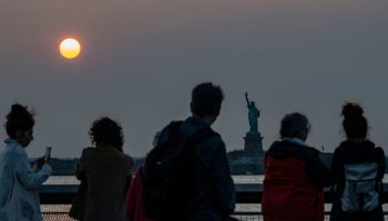People silhouetted by a bright orange setting sun look out over a rail at the Statue of Liberty in the distance.